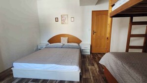 Une chambre avec un lit superposé, un lit simple, un plancher en bois, un mur blanc et une porte en bois.