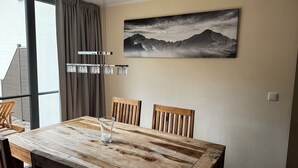 Un comedor con una mesa de madera, cuatro sillas, un florero de vidrio y un cuadro de montañas en la pared.