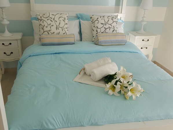 A neatly made bed with a light blue bedspread, two pillows with a floral pattern, a folded white towel, and a bouquet of white flowers. - Kantra Residence (Munxar)