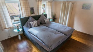 Ein Schlafzimmer mit einem großen Bett, Holzboden, einem kleinen Tisch mit Lampe und einem Fenster mit Vorhängen.