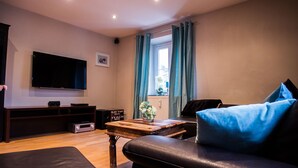 Ein Wohnzimmer mit einem schwarzen Ledersofa, einem Couchtisch aus Holz, einem Flachbildfernseher und einer Lautsprecheranlage.