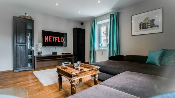 Ein modernes Wohnzimmer mit Couchtisch aus Holz, grauem Sofa und einem Fernseher, der "NETFLIX" zeigt.