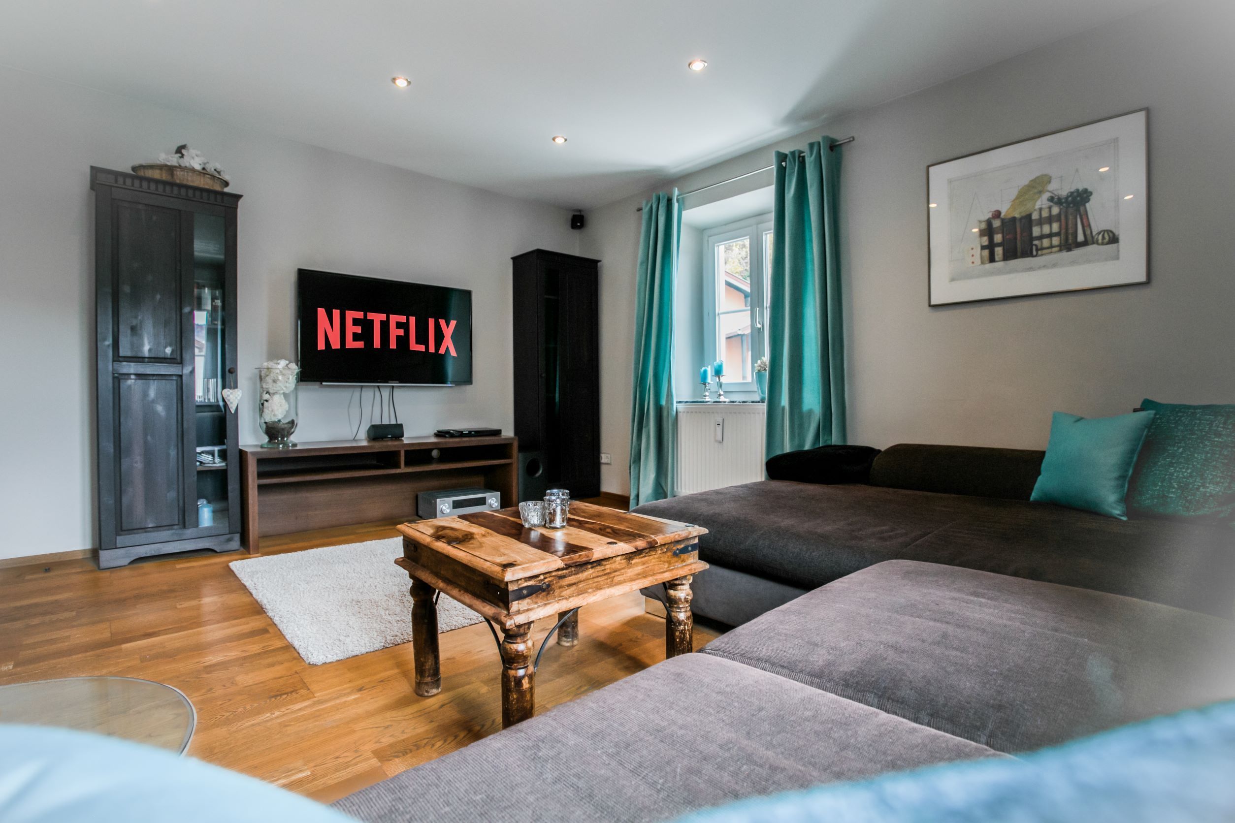 A modern living room with a flat-screen TV displaying 'NETFLIX,' a wooden coffee table, a grey sofa, and a dark wooden cabinet.