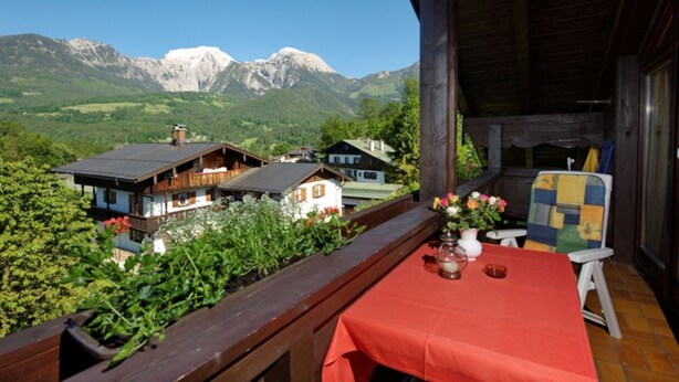 A wooden balcony with a red table and chairs, overlooking a mountain landscape with houses and greenery.