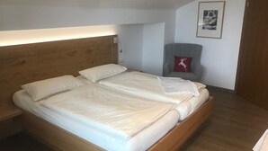 A bedroom with a wooden bed, white bedding, a chair with a red cushion, and a framed picture on the wall.
