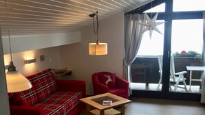 A cozy living area with a red sofa, a wooden coffee table, and a red armchair. There is a large window with curtains and a star decoration.