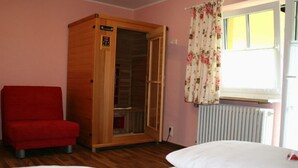 Une pièce avec une armoire en bois, une chaise rouge, un lit avec une literie blanche et une fenêtre ornée de rideaux à motifs floraux.