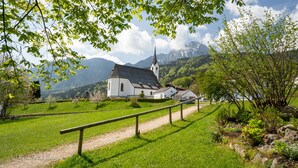 A white church with a steeple, surrounded by lush greenery and mountains in the background. - Ferienwohnung Nr. 5, 48 qm, Nichtraucher, 1 Schlafzimmer, Wohnraum/Kochnische, Balkon, Sat-TV, 4 Sterne, max 3 Personen (Anger)