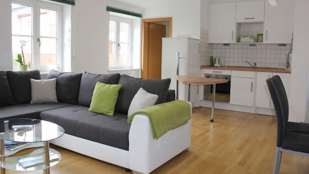 Een moderne woonkamer met een grijze bank, een glazen salontafel en een kleine ronde tafel. Er is een keukenhoek met witte kasten en een houten aanrecht.