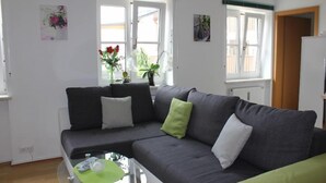 Een moderne woonkamer met een grijze bank, een glazen salontafel en uitzicht buiten door grote ramen.