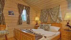 Quarto de cabana de madeira com uma cama, duas mesas de cabeceira, uma cadeira e cortinas.