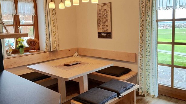 A cozy room with a wooden ceiling, a table with benches, a window with curtains, and a view of a green landscape.