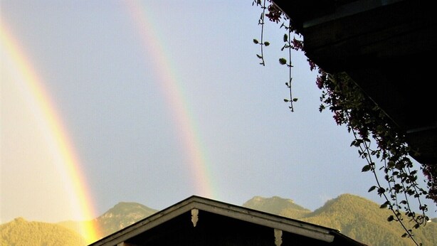 A chalet with a balcony, surrounded by mountains and a rainbow.