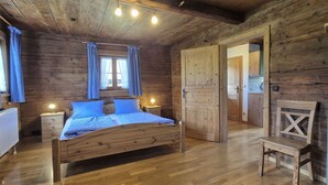 A wooden cabin bedroom with a bed, nightstands, a chair, and a door leading to another room.
