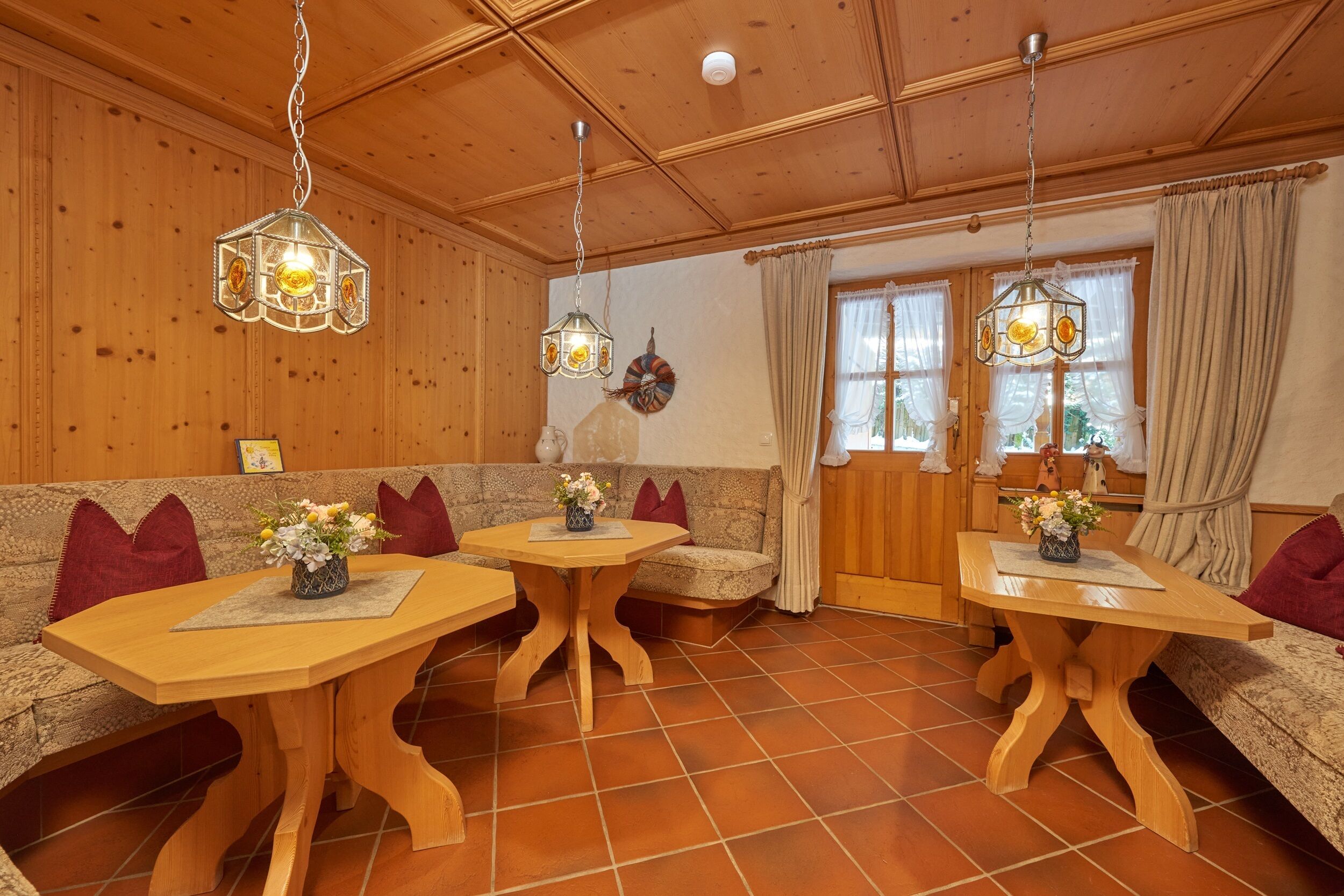 Ein gemütlicher Raum mit Holzwänden und -decke, ausgestattet mit einer Couch, zwei Holztischen mit floralen Mittelstücken und hängenden Pendelleuchten.