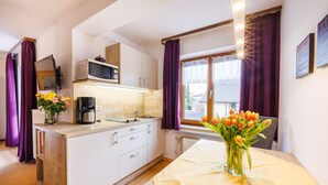 A modern kitchen with white cabinets, a microwave, and a coffee maker. There is a dining table with a yellow tablecloth and a vase of tulips. Purple curtains are visible on the windows. - Gästehaus Wolfgang (Ruhpolding)