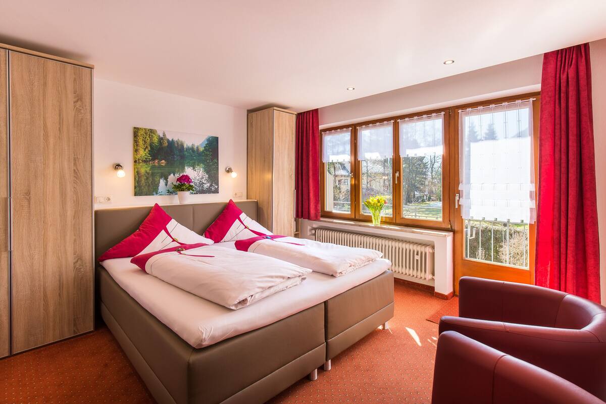 Een slaapkamer met een bed, een stoel, een raam met gordijnen, een radiator en een schilderij aan de muur.