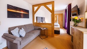 A cozy room with a sofa, a small table, a wooden partition, a bed, a TV, and a window with curtains. - Gästehaus Wolfgang (Ruhpolding)