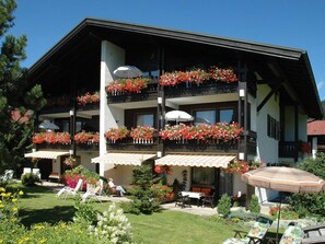 A multi-story hotel with balconies adorned with red flowers, outdoor seating, and a well-maintained garden. - (8) Zwei-Raum-Ferienwohnung 50qm, Bad/WC, Extra-Schlafraum, Küche, Balkon (Reit im Winkl)