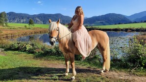 Una mujer vestida monta a caballo junto a un lago, con montañas al fondo.
