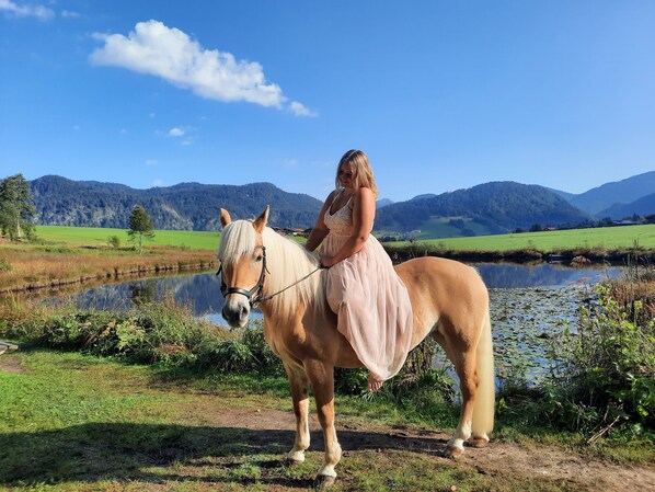 Eine Frau in einem Kleid reitet auf einem Pferd an einem See mit Bergen im Hintergrund.