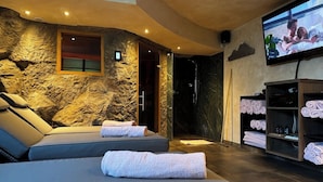A modern spa room with a stone wall, two reclining chairs, a TV, and a shelf with towels. - Alpenglühn (Berchtesgaden)