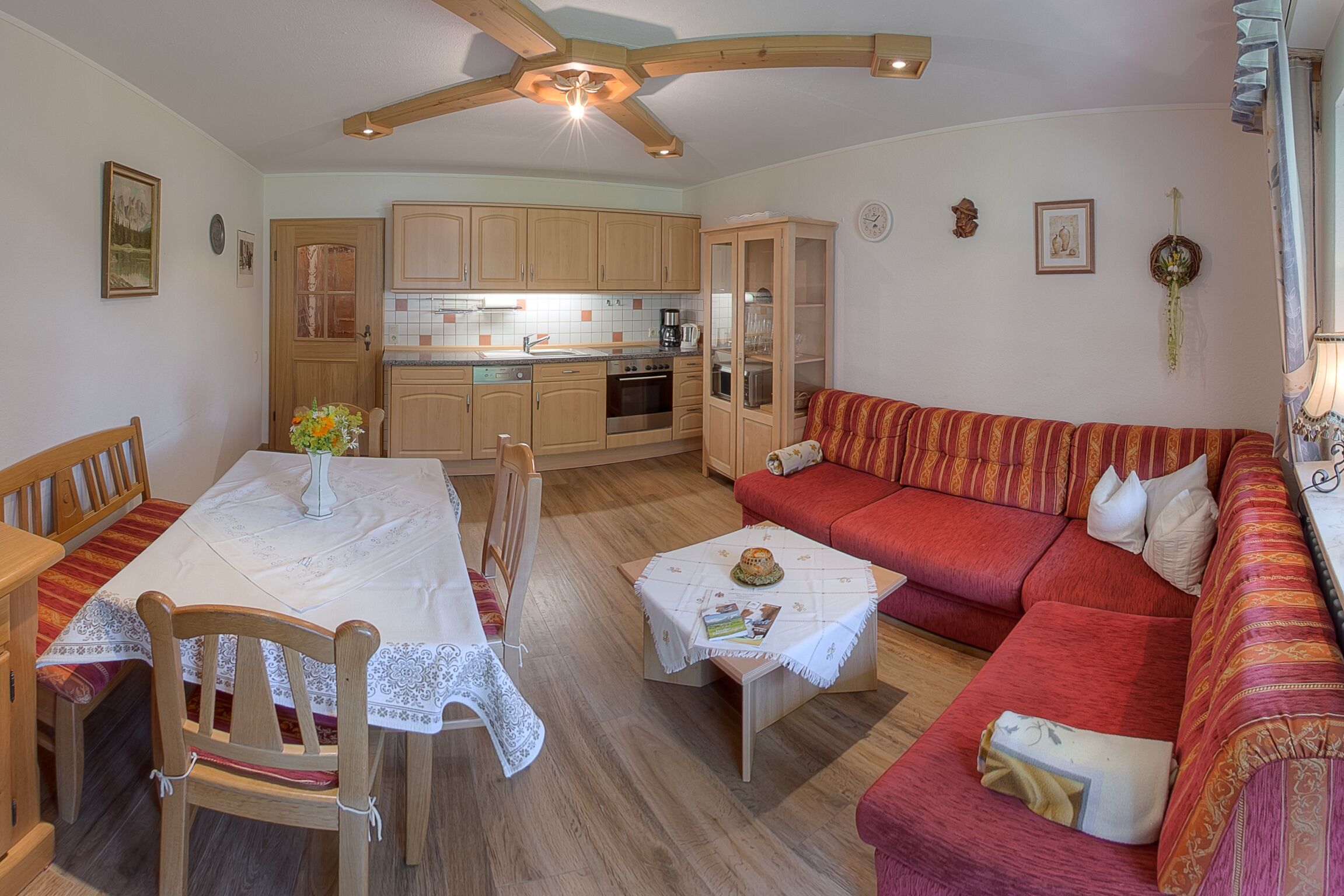A cozy living room with a red sofa, wooden dining table, and chairs. A kitchen with wooden cabinets and a ceiling fan is visible in the background.