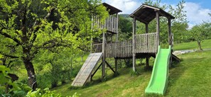 A wooden play structure with a slide, surrounded by lush greenery and trees.