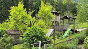 Une structure de jeu en bois avec un toboggan, entourée d’une végétation luxuriante et d’arbres.