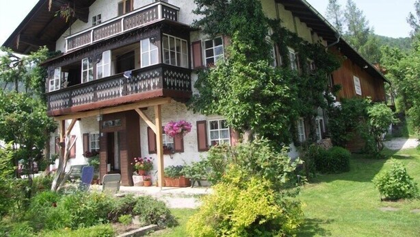 Un chalet de dos pisos con un balcón, rodeado de exuberante vegetación y un jardín cuidado.