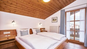 A double bed with a wooden headboard, two bedside tables, a nightstand, a radiator, and a window with curtains. - (5) Two-room vacation apartment, approx. 42 m², separate kitchen and balcony (Reit im Winkl)