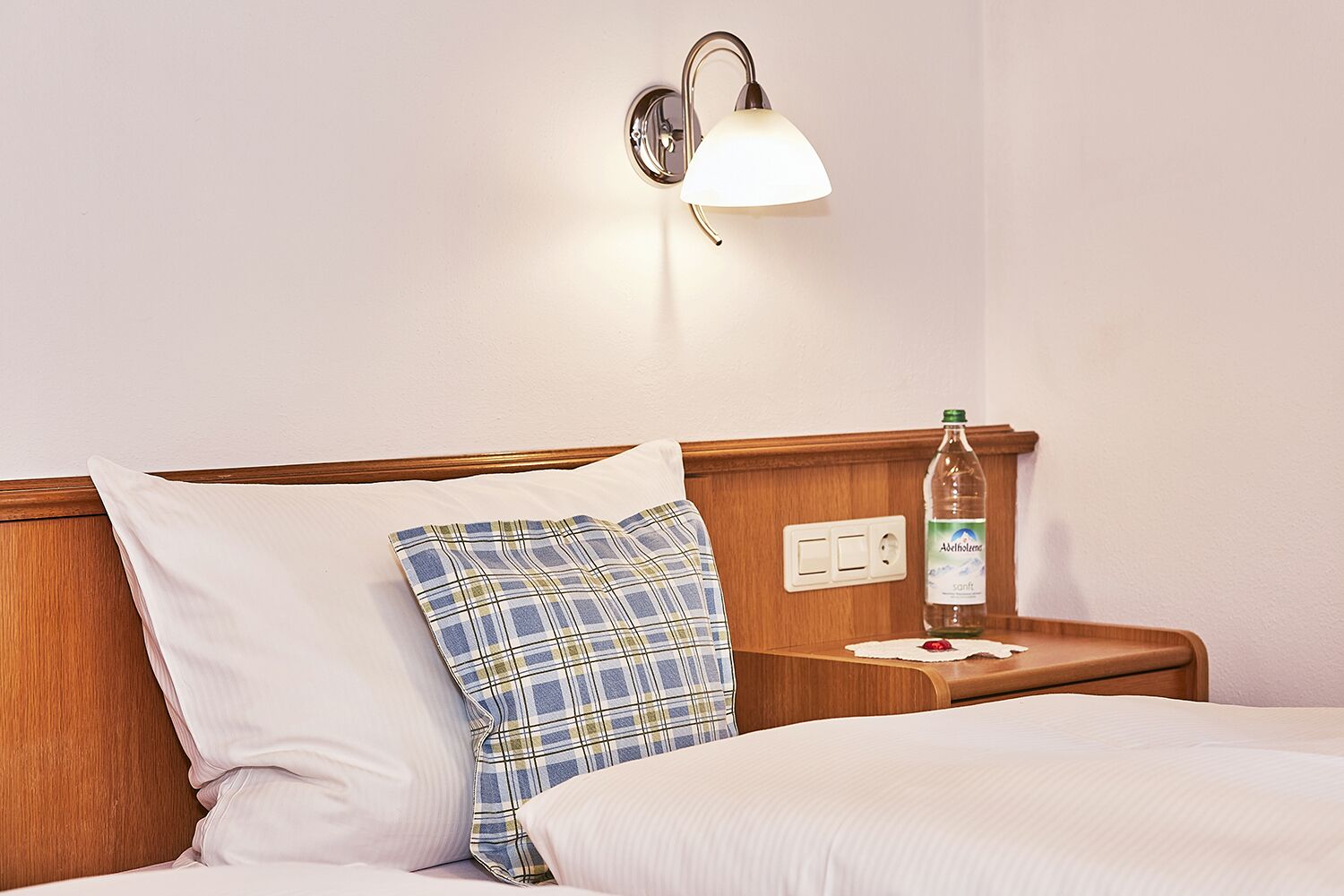 A hotel room with a bed, a nightstand, a wall-mounted lamp, a bottle of water, and a power outlet.
