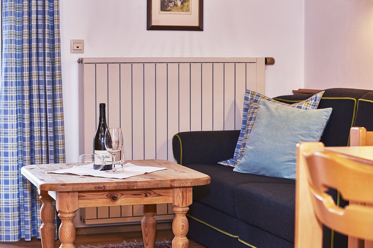 A cozy living room with a plaid curtain, a wooden table with a bottle and glass, a black sofa with blue pillows, and a framed picture on the wall.