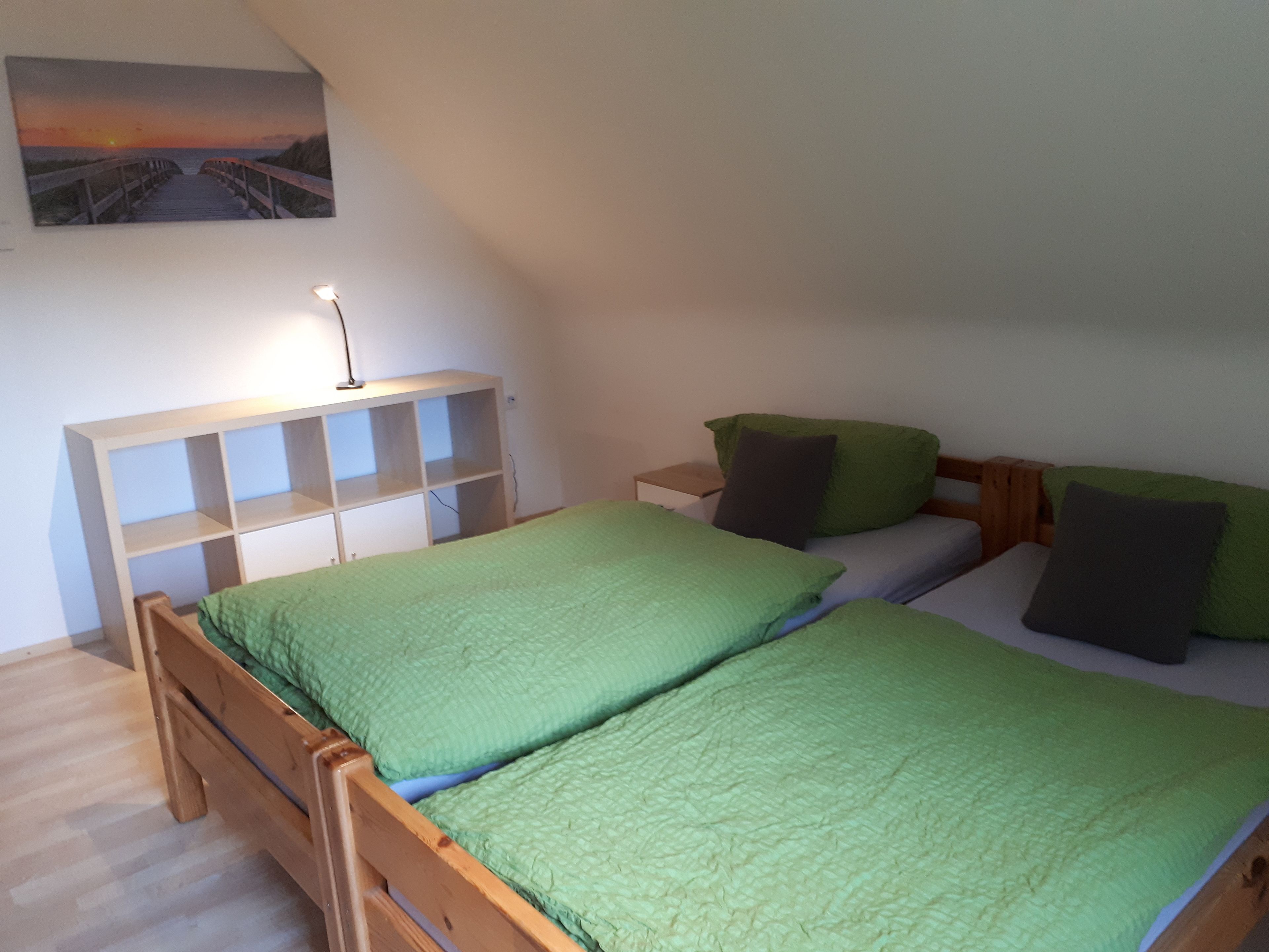 Une chambre à coucher avec un lit en bois, une literie verte, une table de chevet blanche, une lampe et un tableau représentant un coucher de soleil accroché au mur.