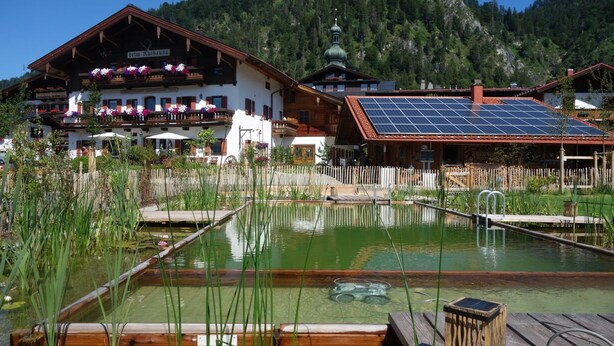 A traditional Alpine building with a wooden facade, a solar panel roof, and a pond in the foreground. - Ferienwohnung Erdgeschoß (Reit im Winkl)
