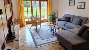 Un salon avec un canapé gris, un fauteuil en osier, une table basse, une plante en pot et une grande fenêtre ornée de rideaux jaunes.