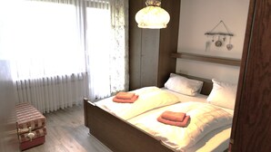 A bedroom with a wooden bed, white bedding, a hanging light fixture, a wall-mounted shelf with decorative items, and a window with sheer curtains. - (5) Zwei-Raum-Ferienwohnung 50qm, Bad/WC, Extra-Schlafraum, Küche, Balkon (Reit im Winkl)