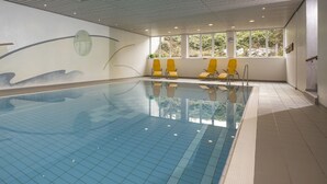 An indoor swimming pool with clear blue water, yellow chairs along the side, and a view of greenery through large windows.