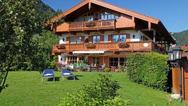 A two-story chalet with a balcony, a garden with chairs, and a mountain backdrop. - One-room apartment 30sqm, shower/WC, kitchen, balcony (Reit im Winkl)