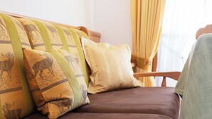 A brown sofa with patterned cushions and a wooden chair in a room with a window and curtains. - One-room apartment 30sqm, shower/WC, kitchen, balcony (Reit im Winkl)