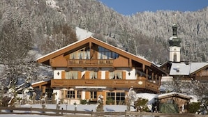 A large wooden chalet with a balcony, surrounded by snow-covered trees and a mountain backdrop. - One-room apartment 30sqm, shower/WC, kitchen, balcony (Reit im Winkl)