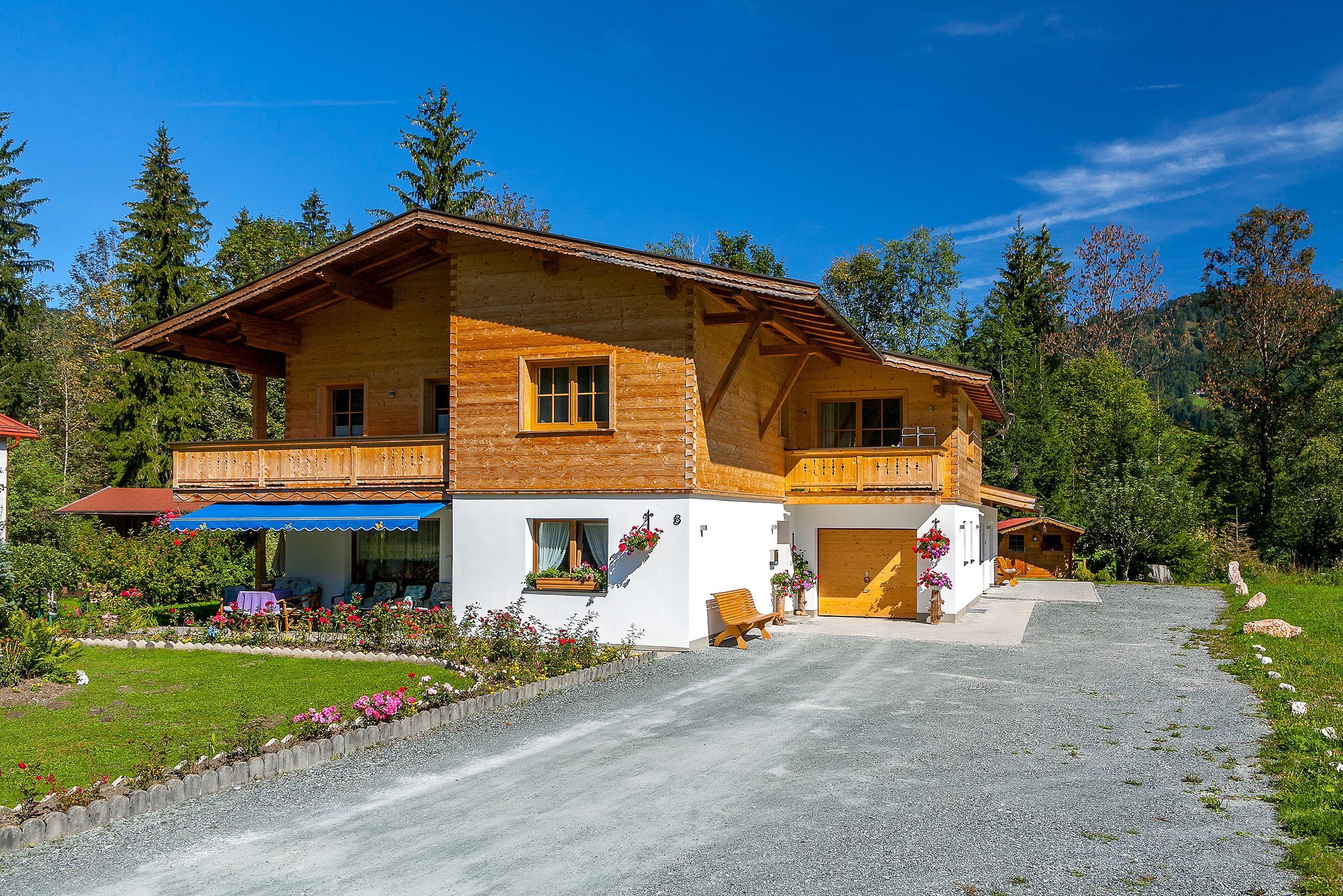 Une maison en bois à deux étages, dotée d’un balcon, d’une allée en gravier et d’un jardin fleuri.