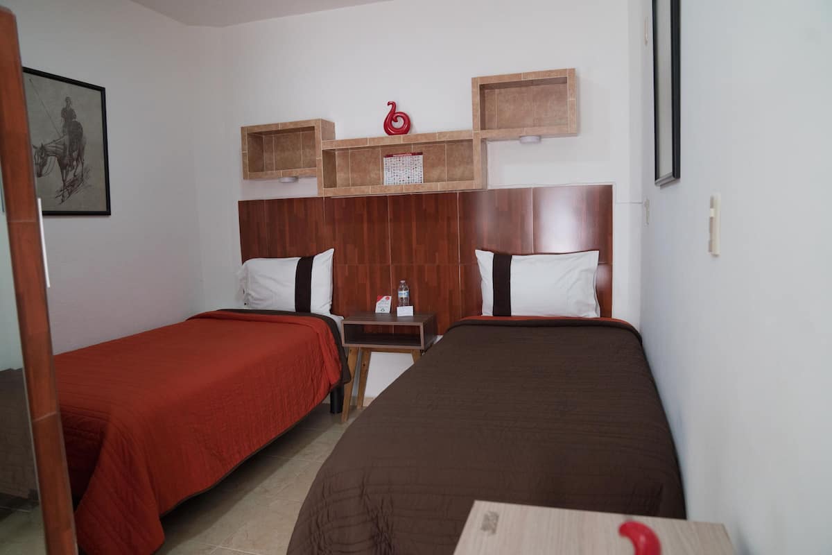 Habitación de hotel con dos camas individuales, cabeceros de madera, una mesita con una botella de agua y un cuadro enmarcado en la pared.
