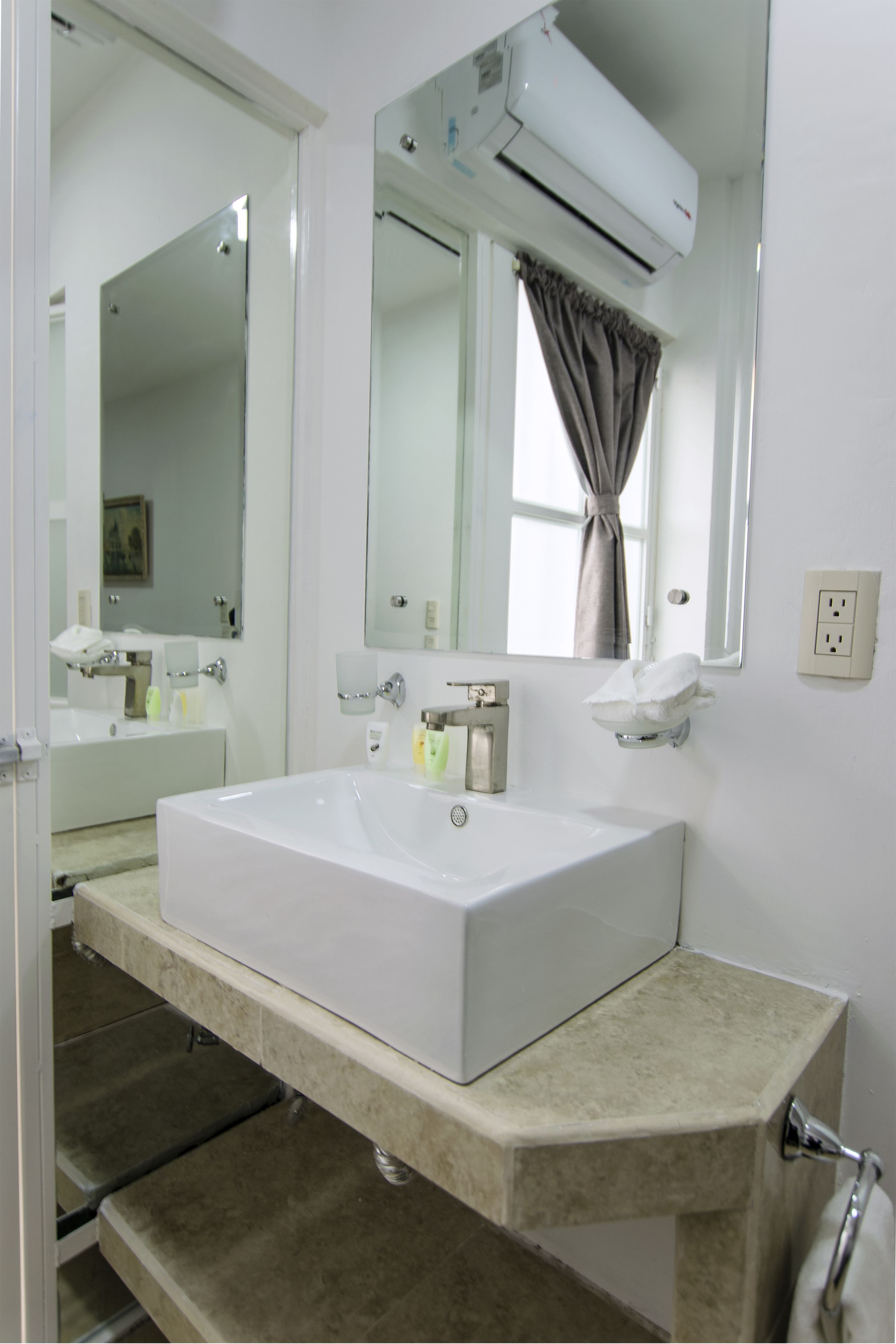 Un baño con lavamanos blanco, espejo y equipo de aire acondicionado.