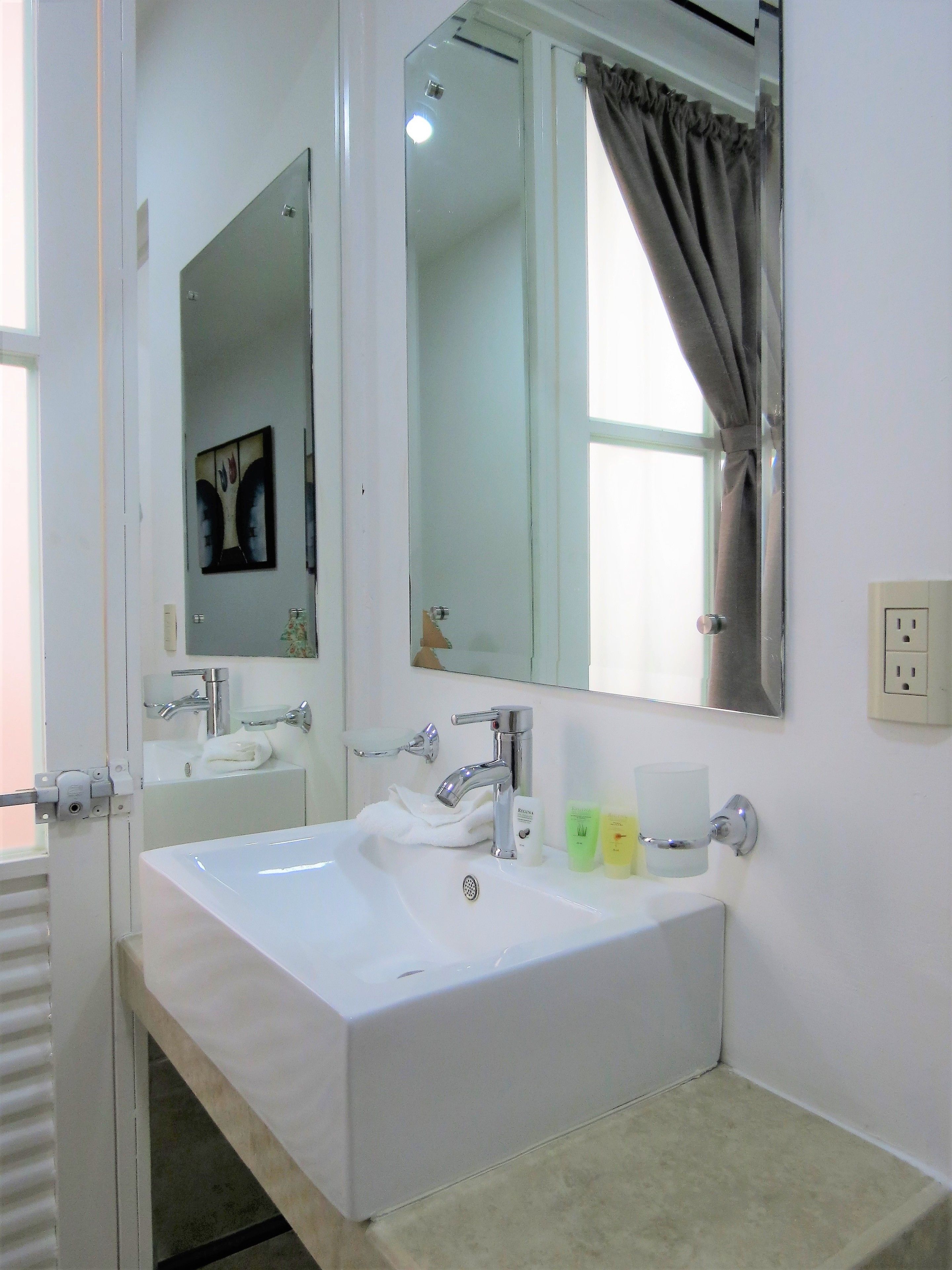 Un baño con un lavamanos blanco, dos espejos, una ventana con cortinas y un perchero para toallas.