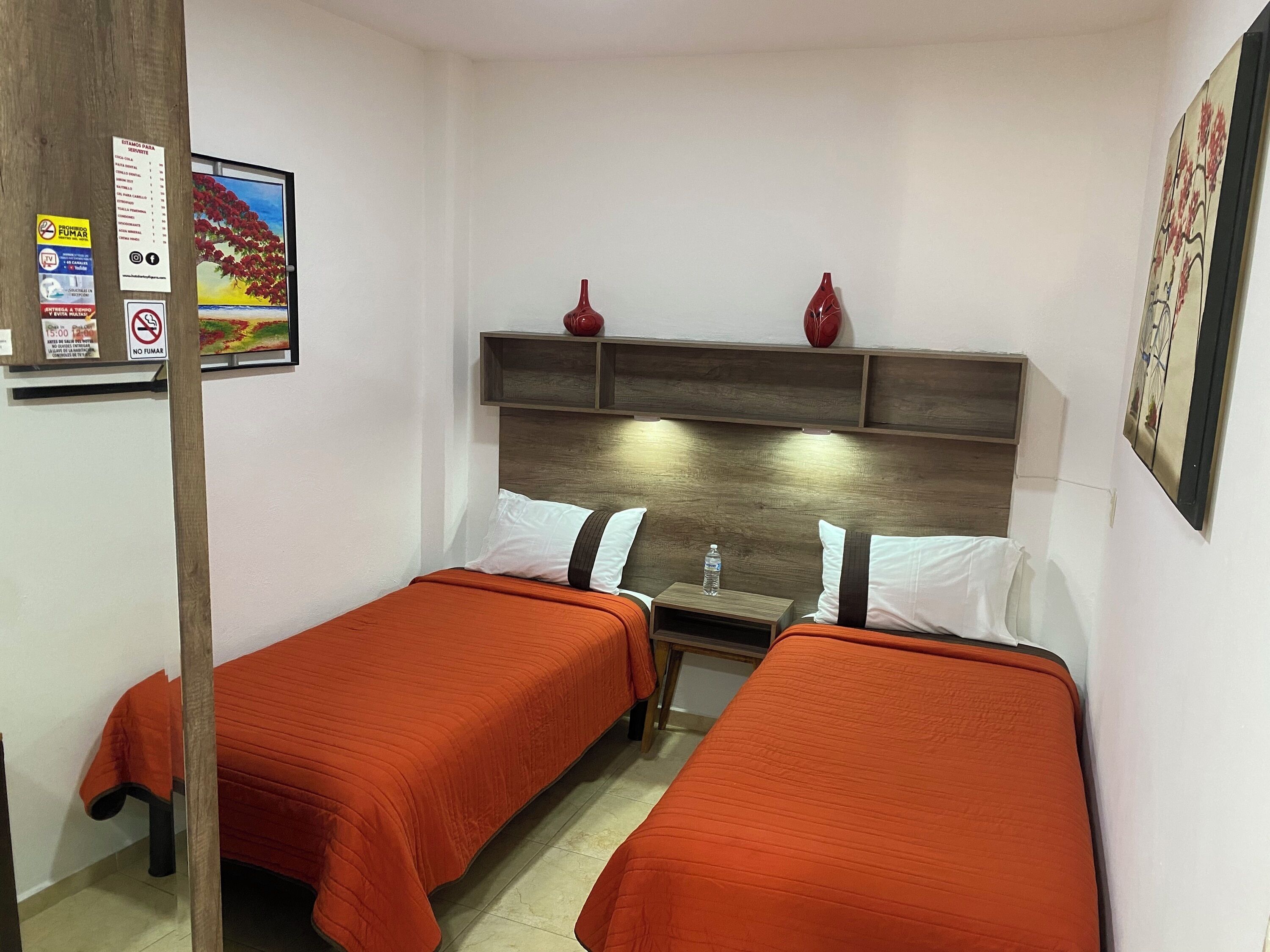 Een hotelkamer met twee bedden, elk met een oranje sprei, een houten hoofdbord en een klein tafeltje met een waterfles. Op het hoofdbord staan twee rode vazen.