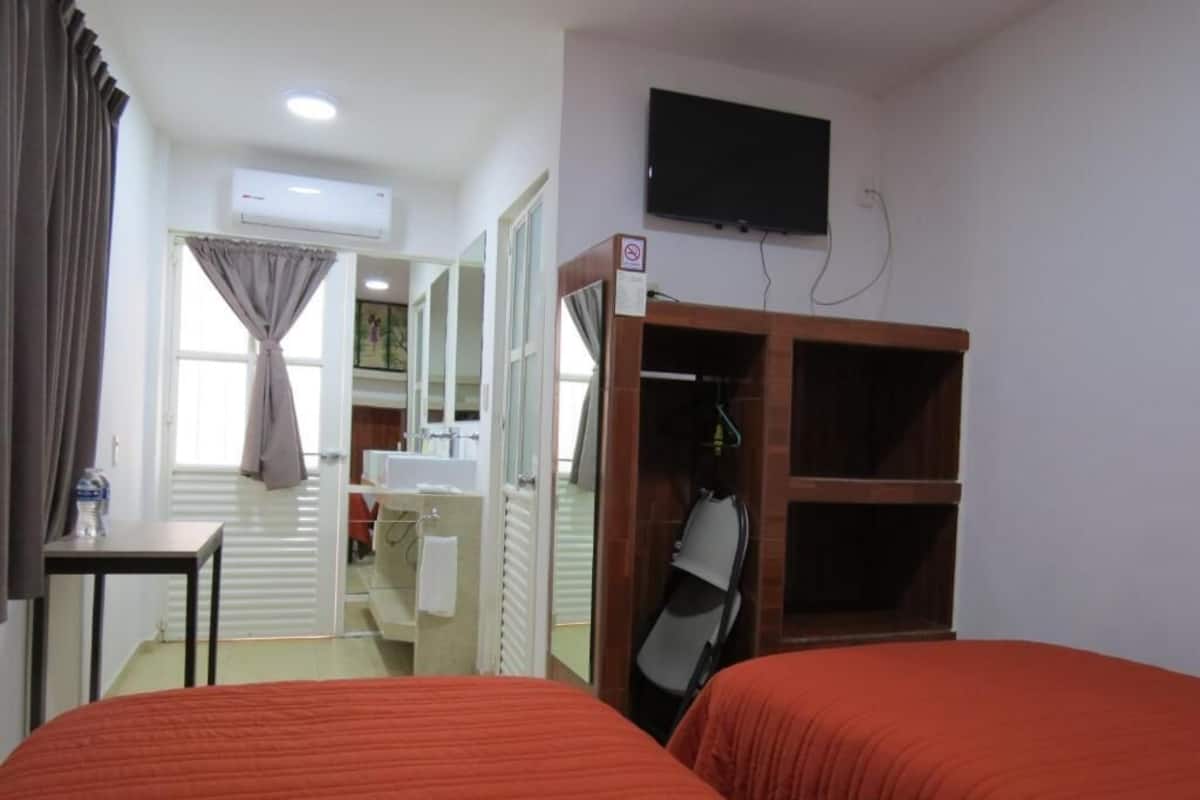 Habitación de hotel con dos camas, televisión, aire acondicionado, perchero y baño visible a través de una puerta abierta.