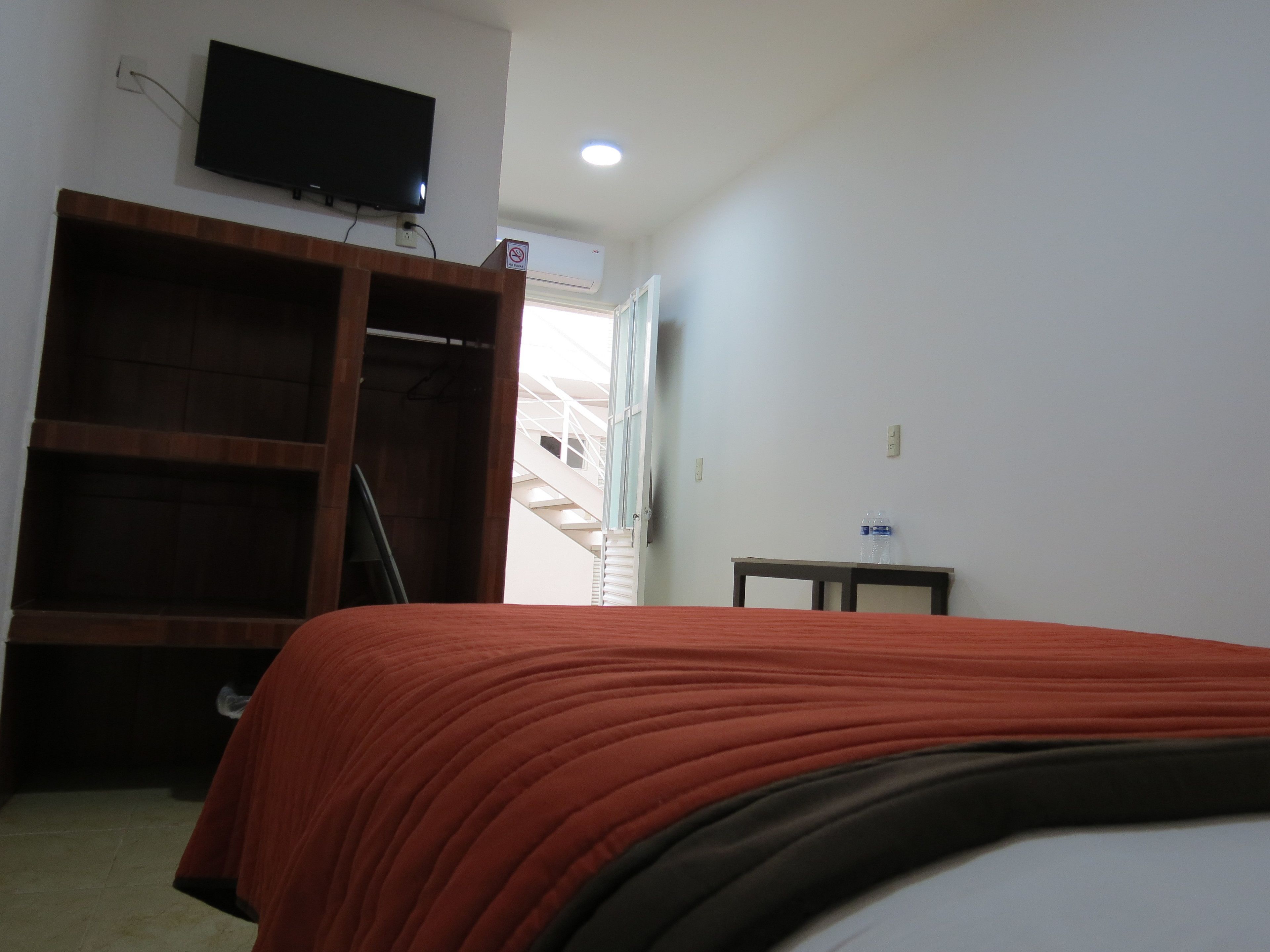 A bedroom with a bed, a TV mounted on the wall, a wooden wardrobe, and a small table with a water bottle.