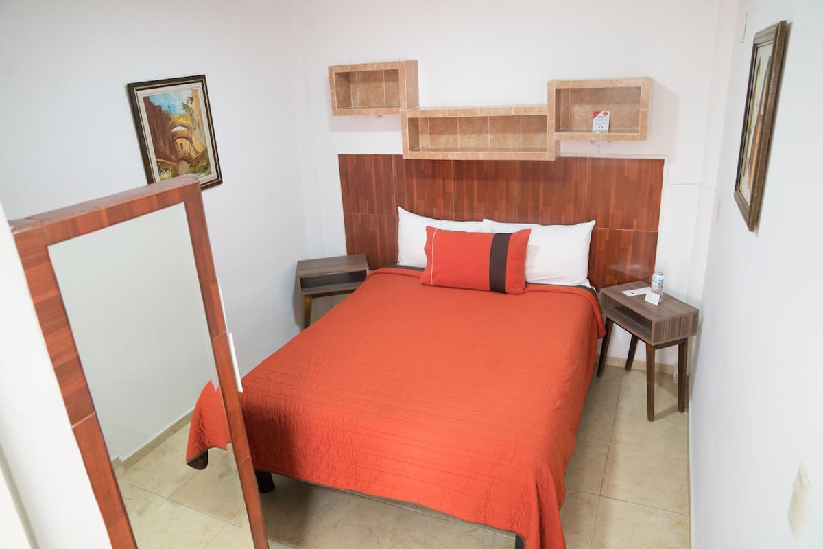 Un dormitorio con cabecera de madera, colcha roja, dos mesitas de noche y una pequeña estantería sobre la cama.