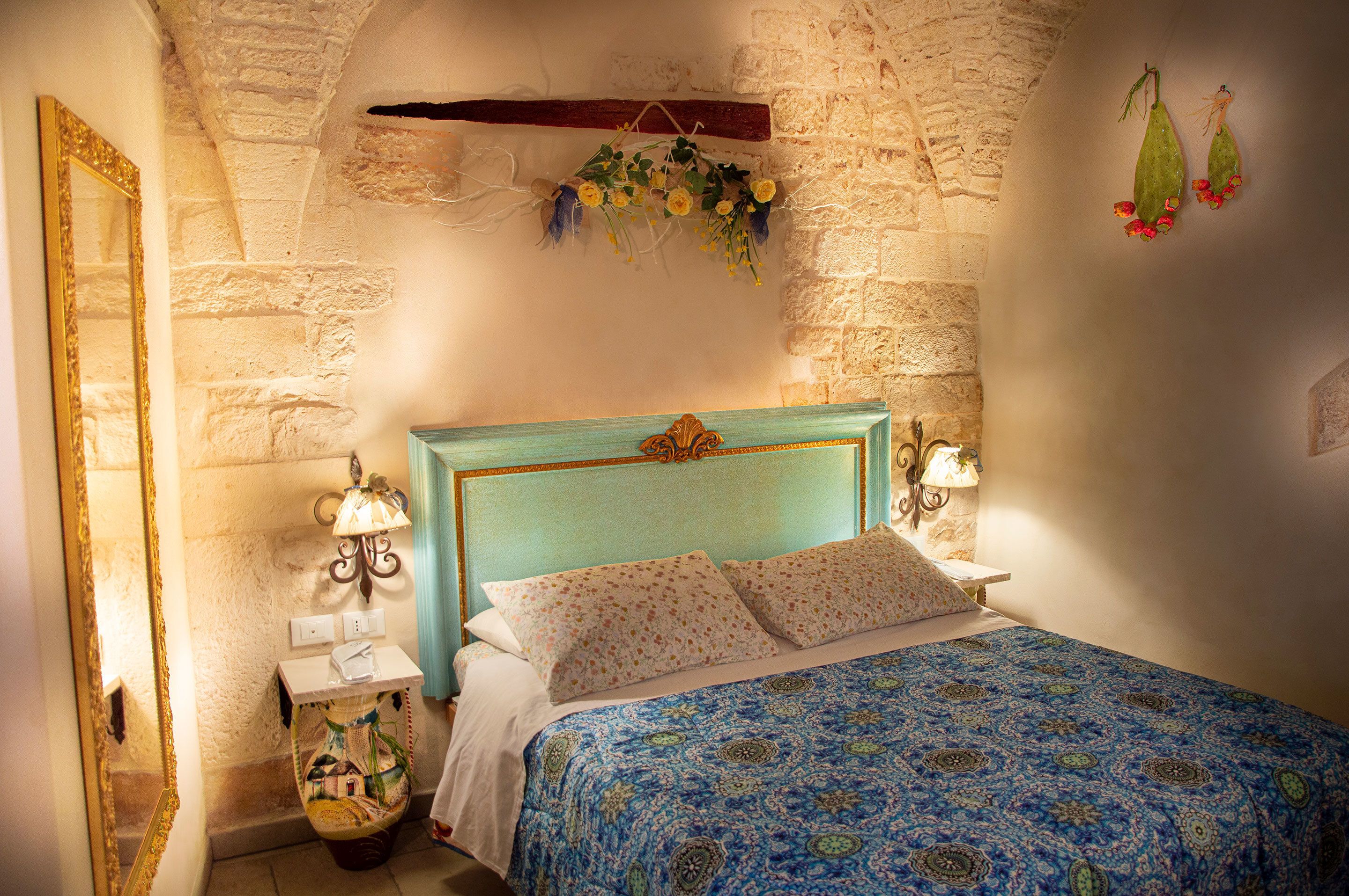 A bedroom with a stone wall, a bed with a blue patterned bedspread, a wooden headboard, and a hanging floral decoration.